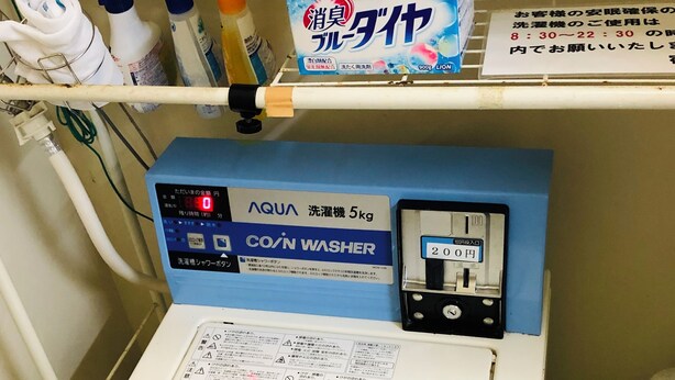 A laundry machine with a control panel, a box of laundry detergent, and cleaning supplies on a shelf.