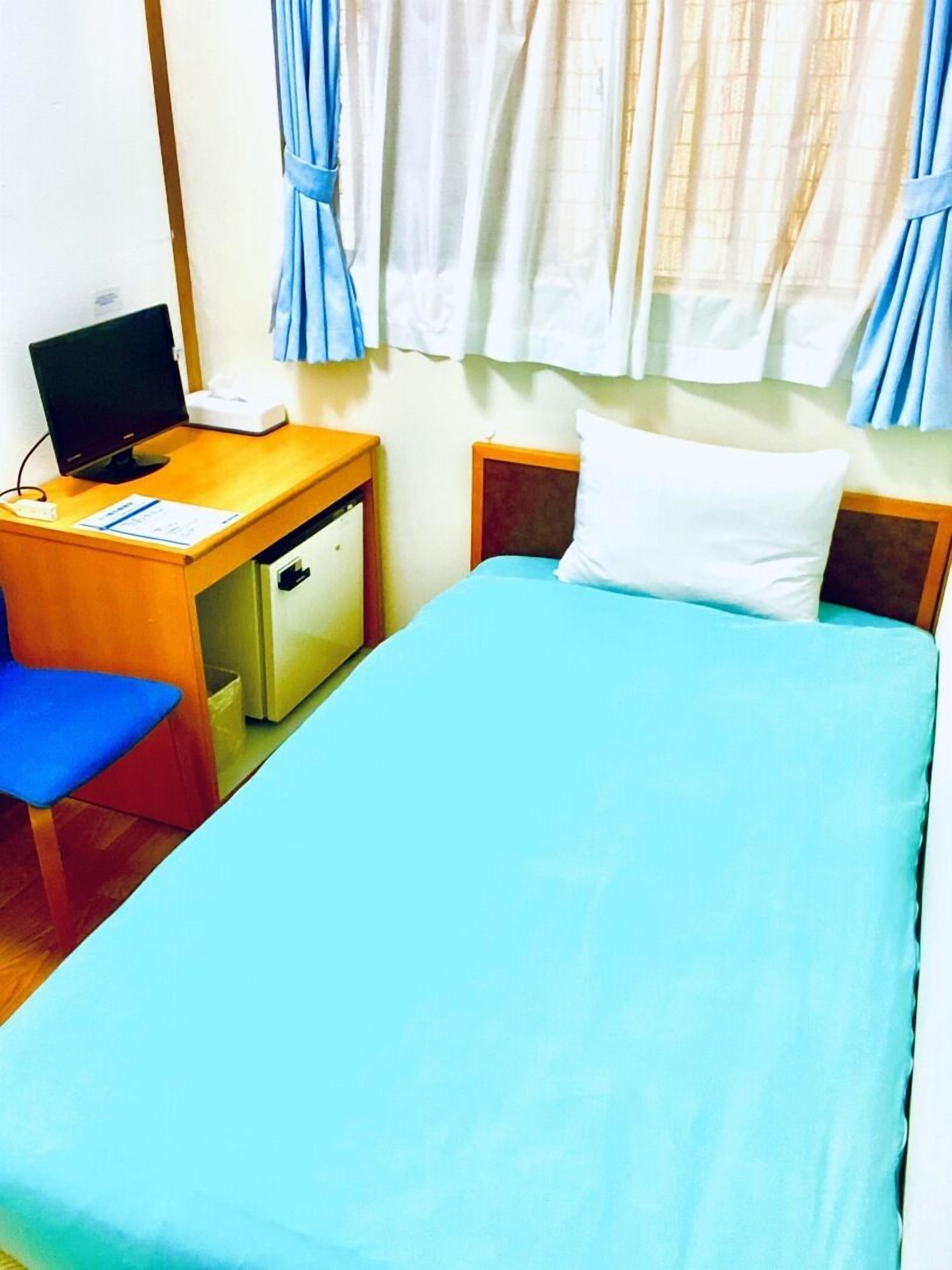Ein Einzelbett mit blauer Matratze und weißem Kopfkissen, ein Schreibtisch mit Computer, ein Stuhl und eine kleine Kommode.