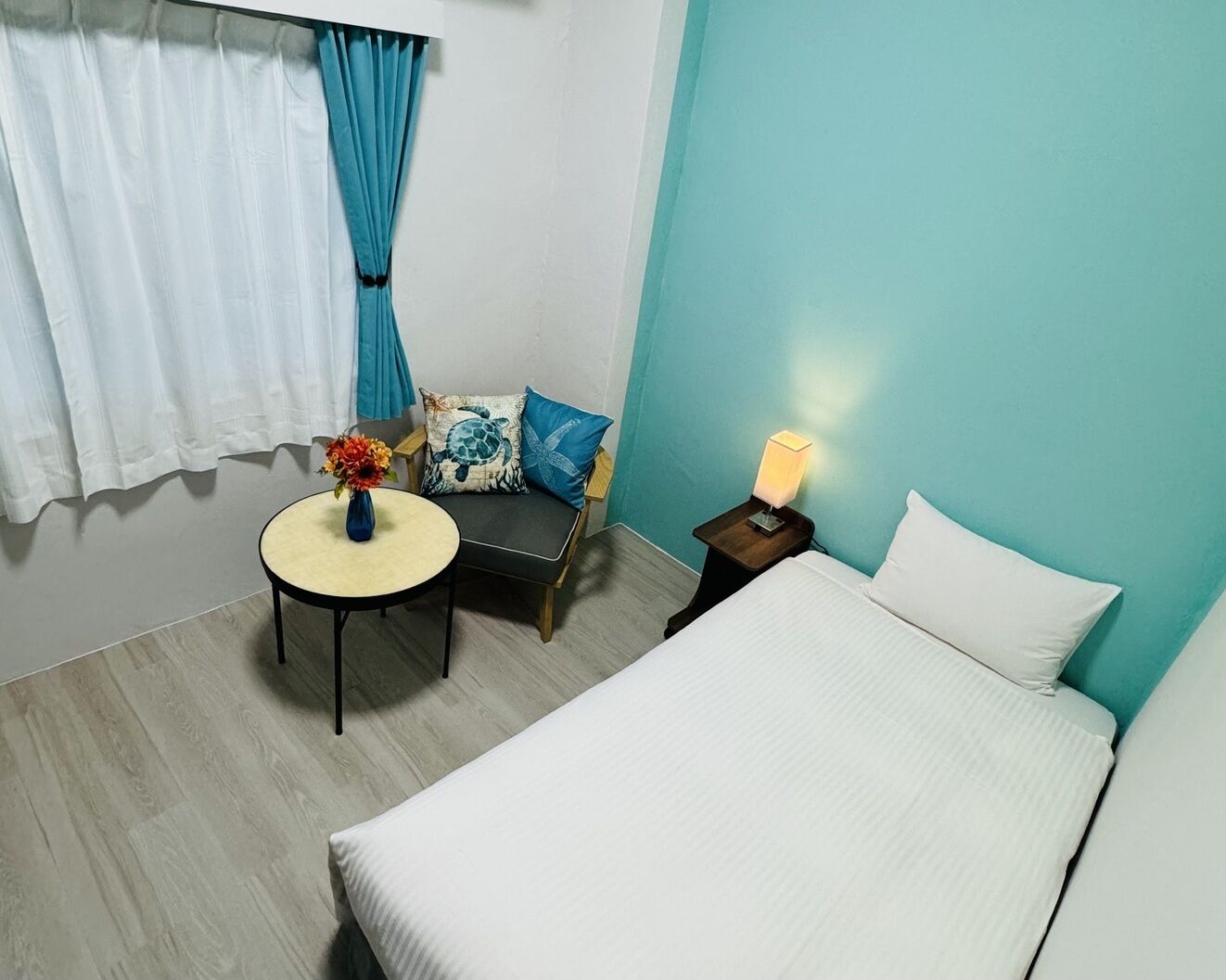 Ein Hotelzimmer mit einem Bett, einem kleinen runden Tisch mit Blumen in einer Vase, einem Sessel mit Kissen und einem Nachttisch mit einer Lampe.