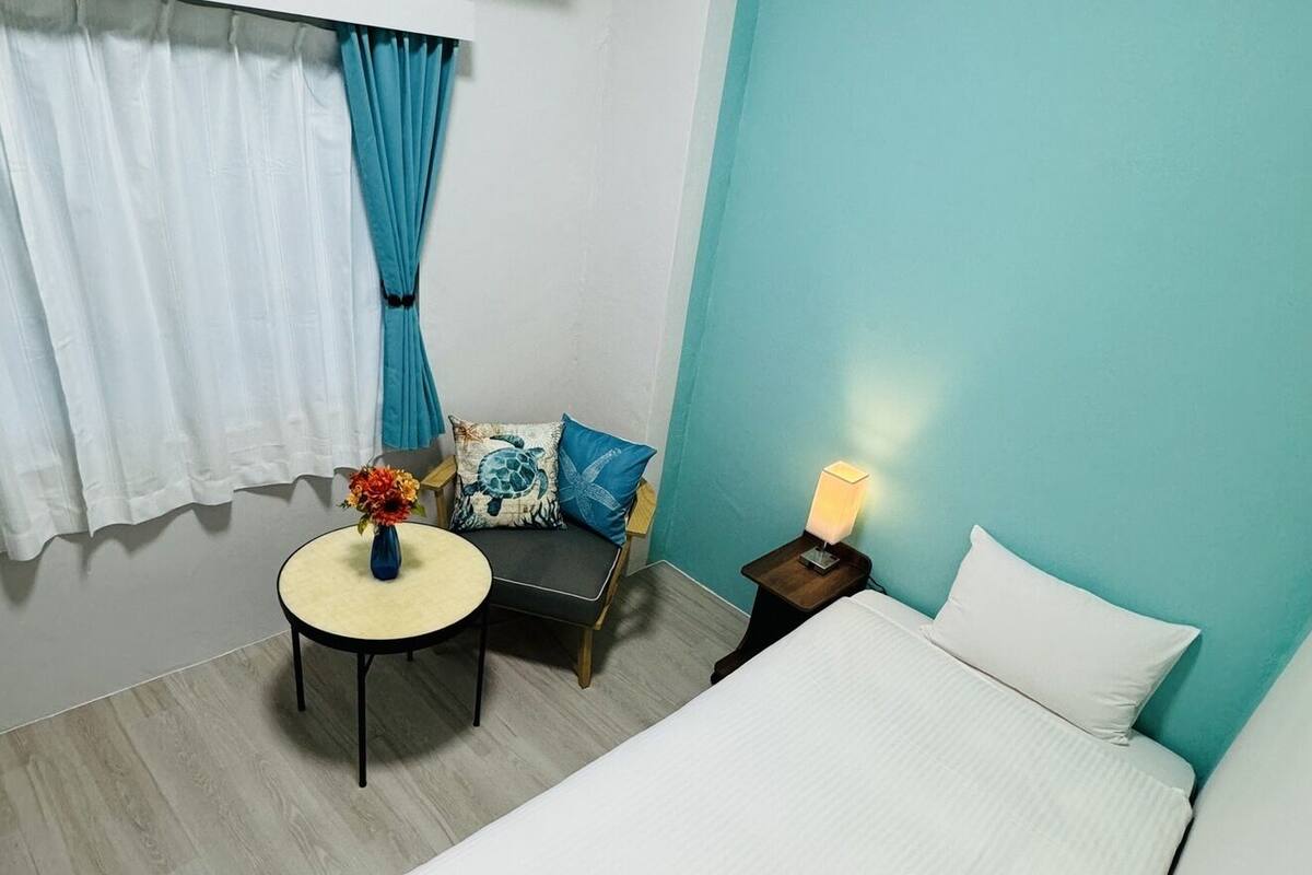 Ein Hotelzimmer mit einem Bett, einem kleinen runden Tisch mit Blumen in einer Vase, einem Sessel mit Kissen und einem Nachttisch mit einer Lampe.