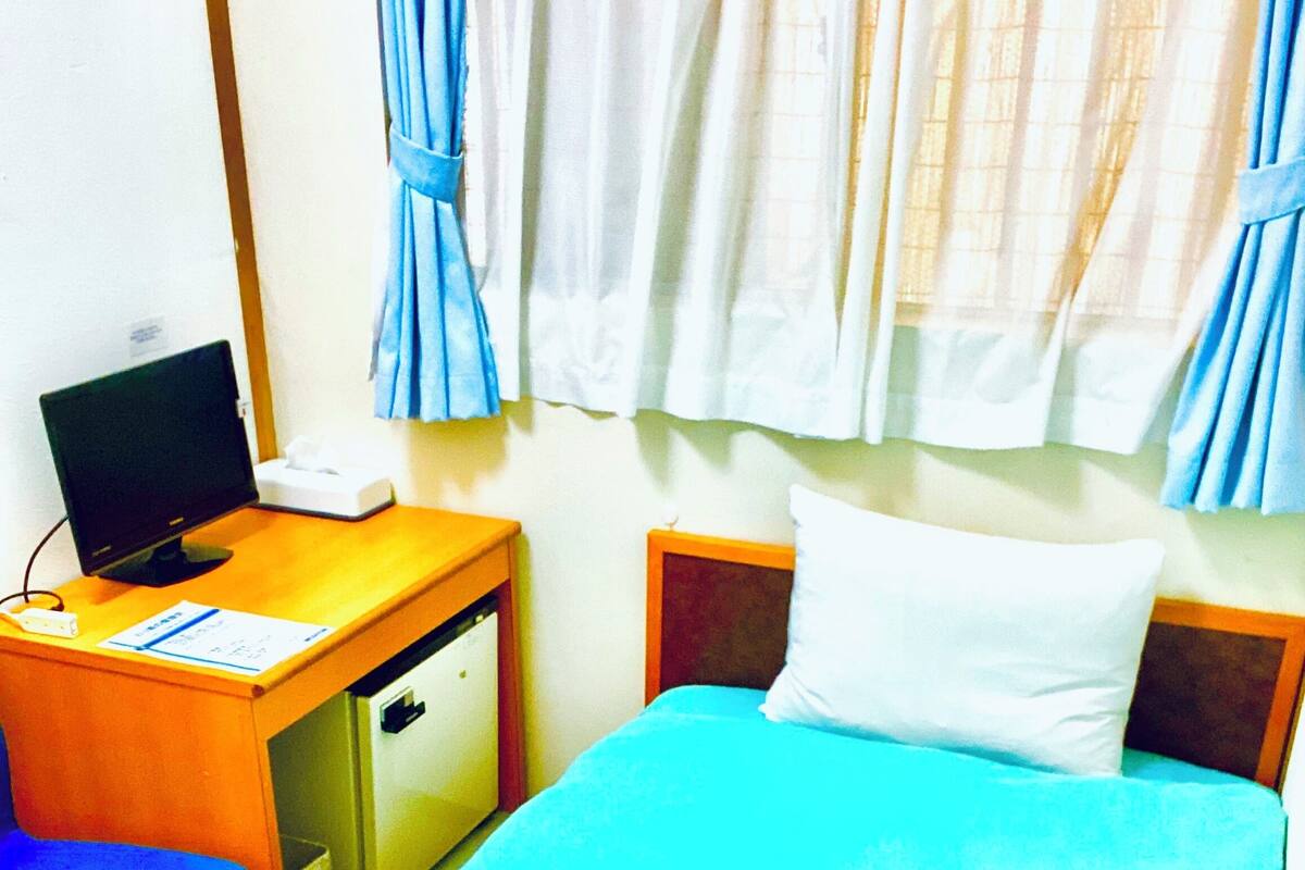 Ein Zimmer mit einem Bett, einem Schreibtisch mit Computer, einem Stuhl und einem kleinen Kühlschrank.