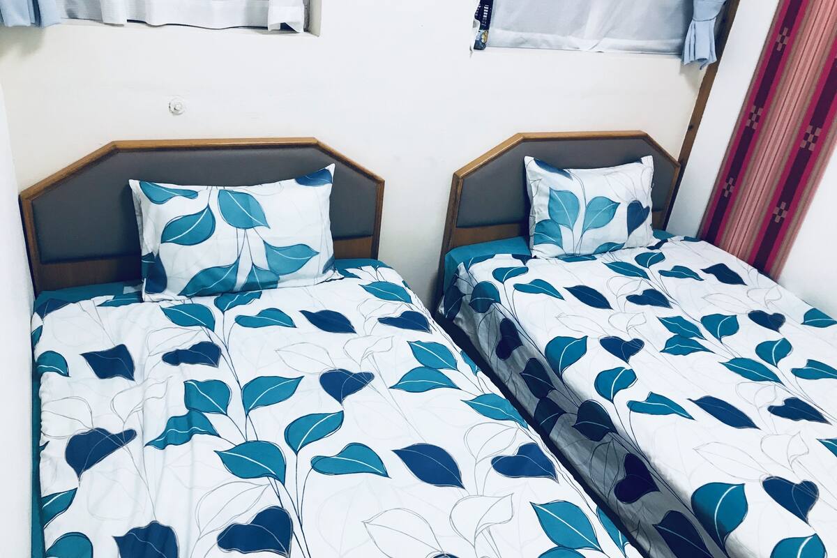 Zwei Doppelbetten mit blattgemusterten Bettbezügen, Holz-Kopfteile und eine schlichte Wand.