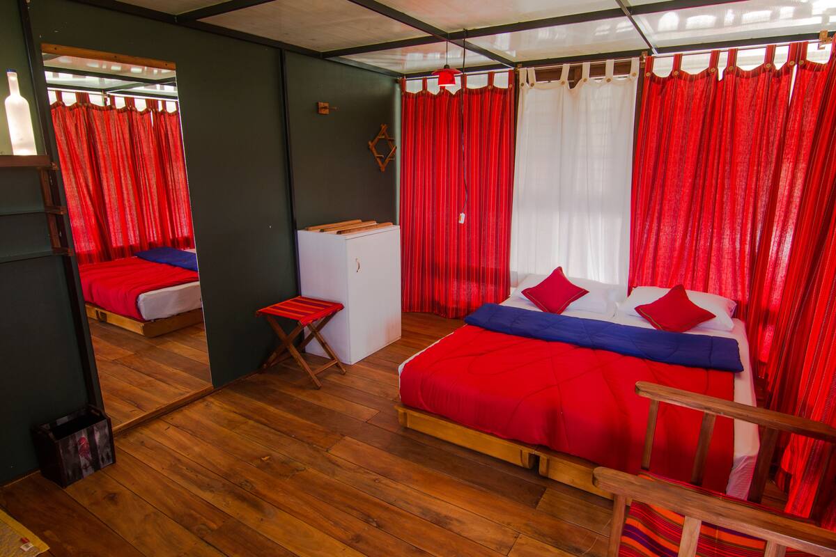 Una habitación con piso de madera, una cama con ropa de cama roja y azul, una pequeña mesa de madera y un espejo en la pared.