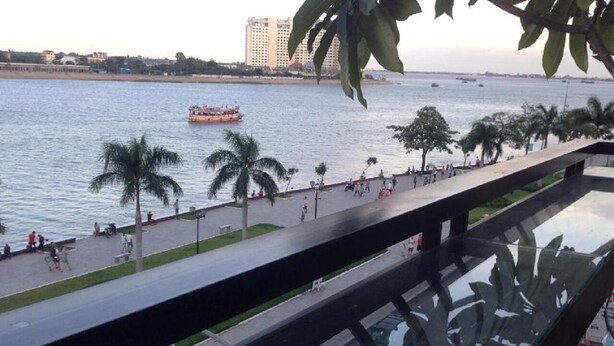 A balcony with a glass railing overlooking a waterfront promenade with palm trees and a boat in the distance. - New Season Riverside Hotel (Phnom Penh)