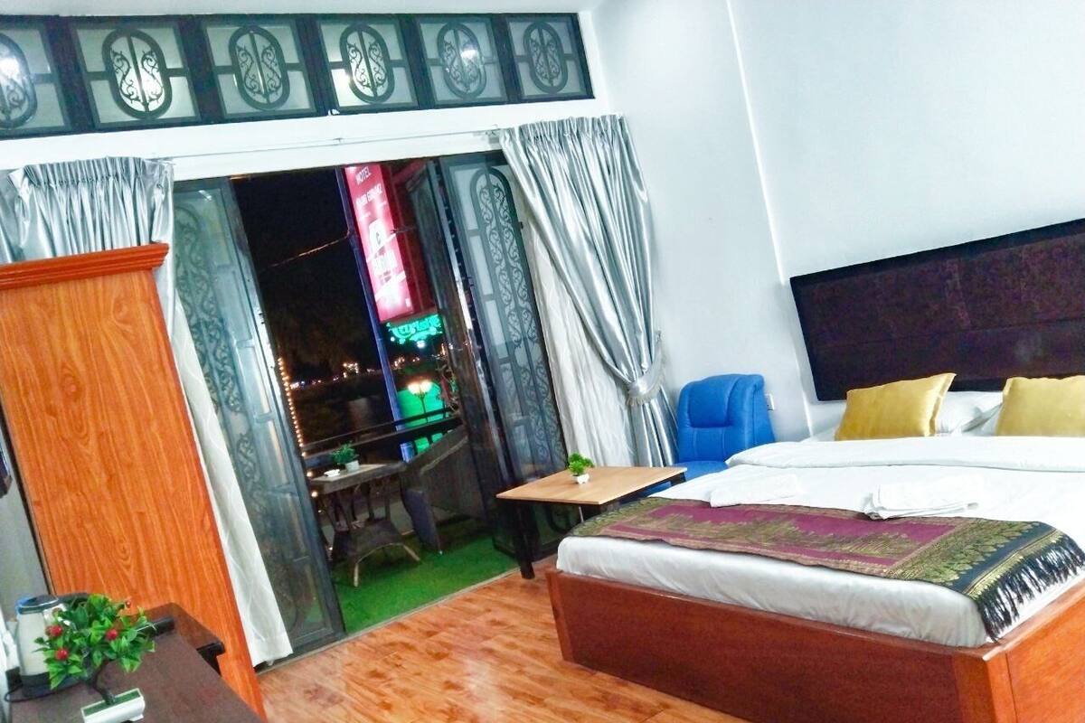 Habitación de hotel con suelo de madera, cama con cubrecama blanco y rojo, una mesita con una planta y vista a un paisaje urbano a través de una puerta de vidrio.
