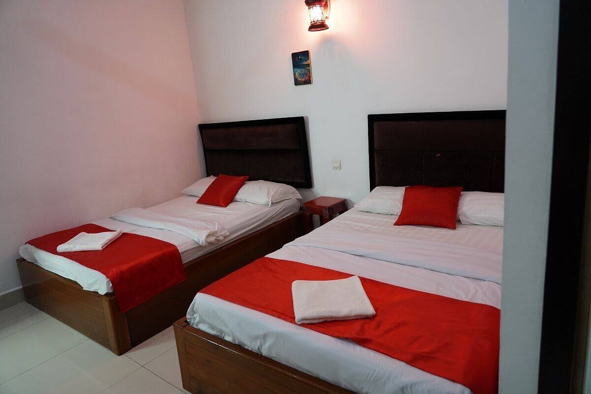 Dos camas individuales con cabeceros de madera y ropa de cama roja, una luz nocturna y un pequeño cuadro en la pared.