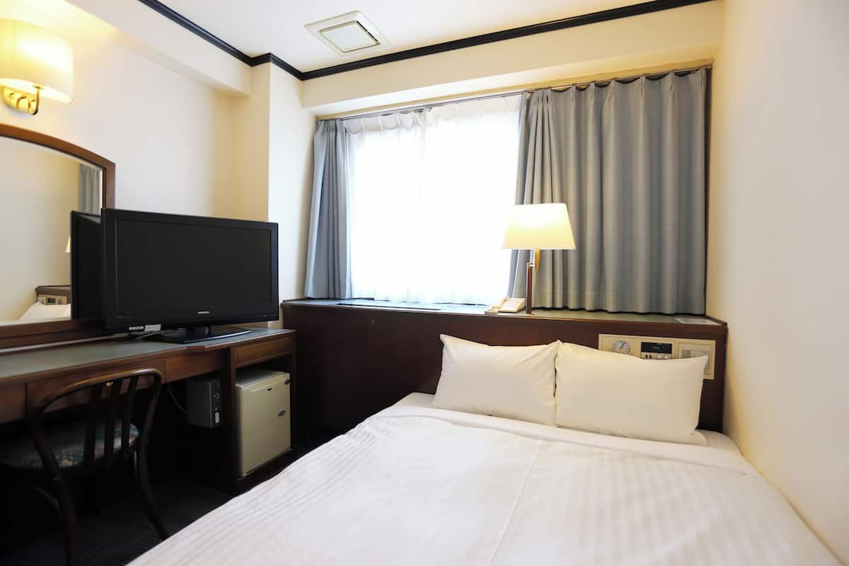 A hotel room with a bed, a desk with a television, a chair, a lamp, and a window with curtains.
