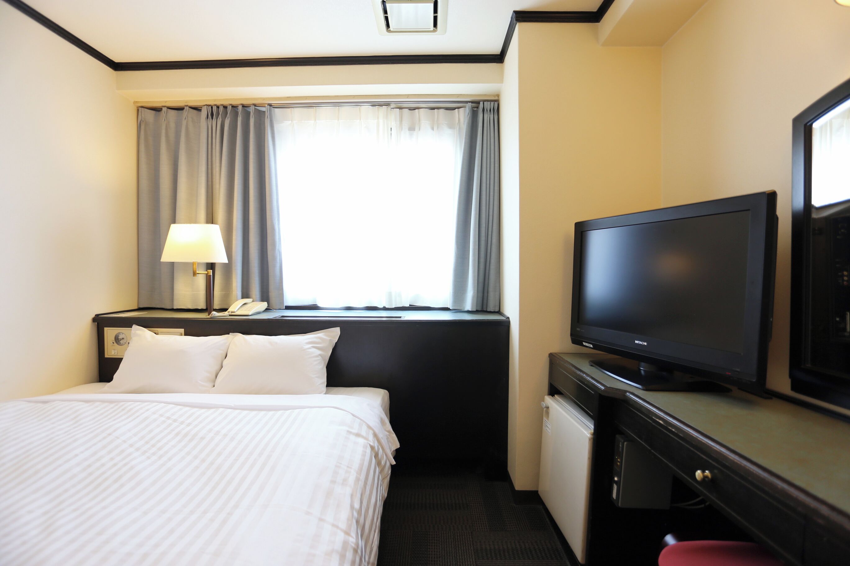 A hotel room with a bed, a desk with a television, a lamp, and a window with curtains.