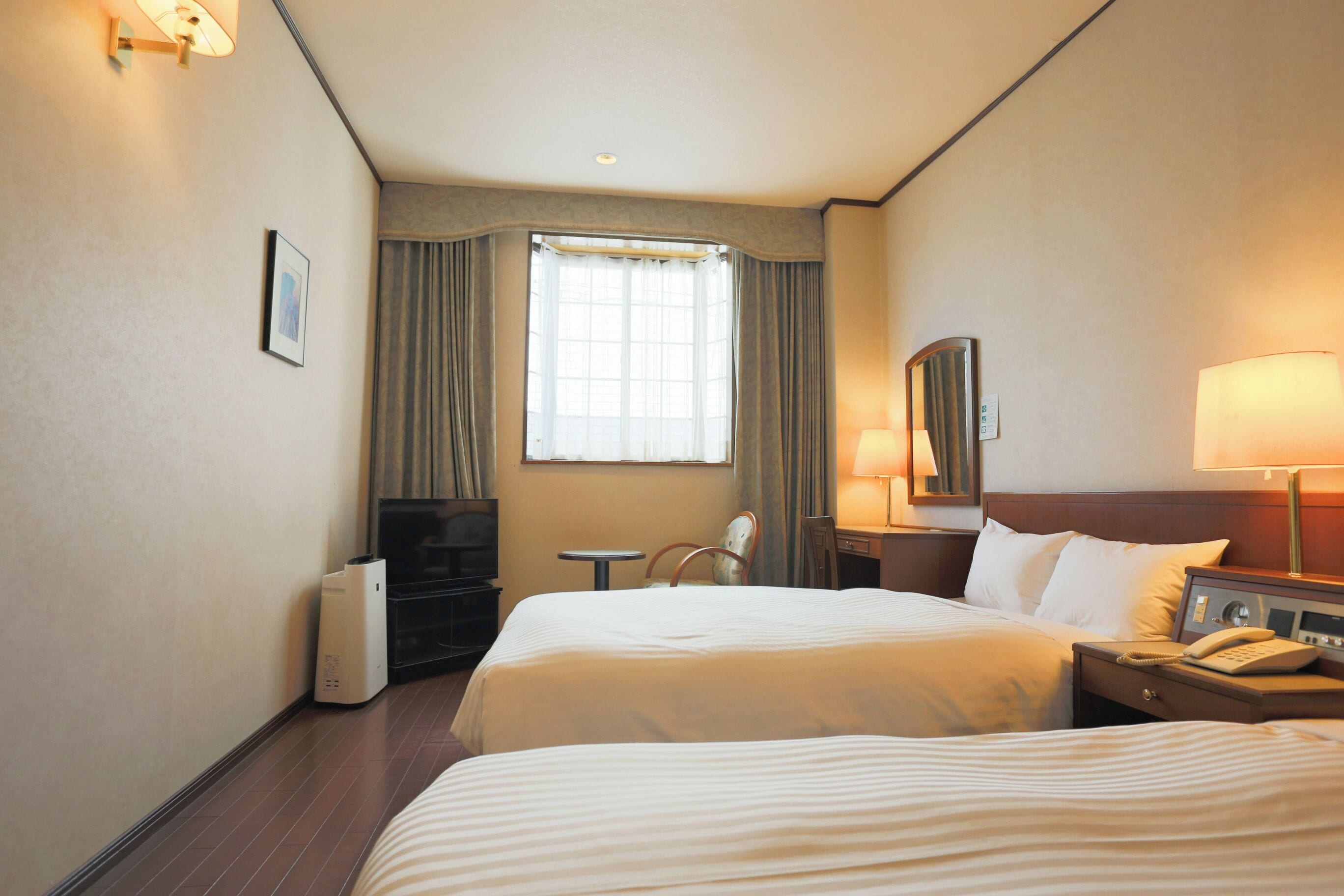 A hotel room with two beds, a desk, a chair, a TV, and a lamp.