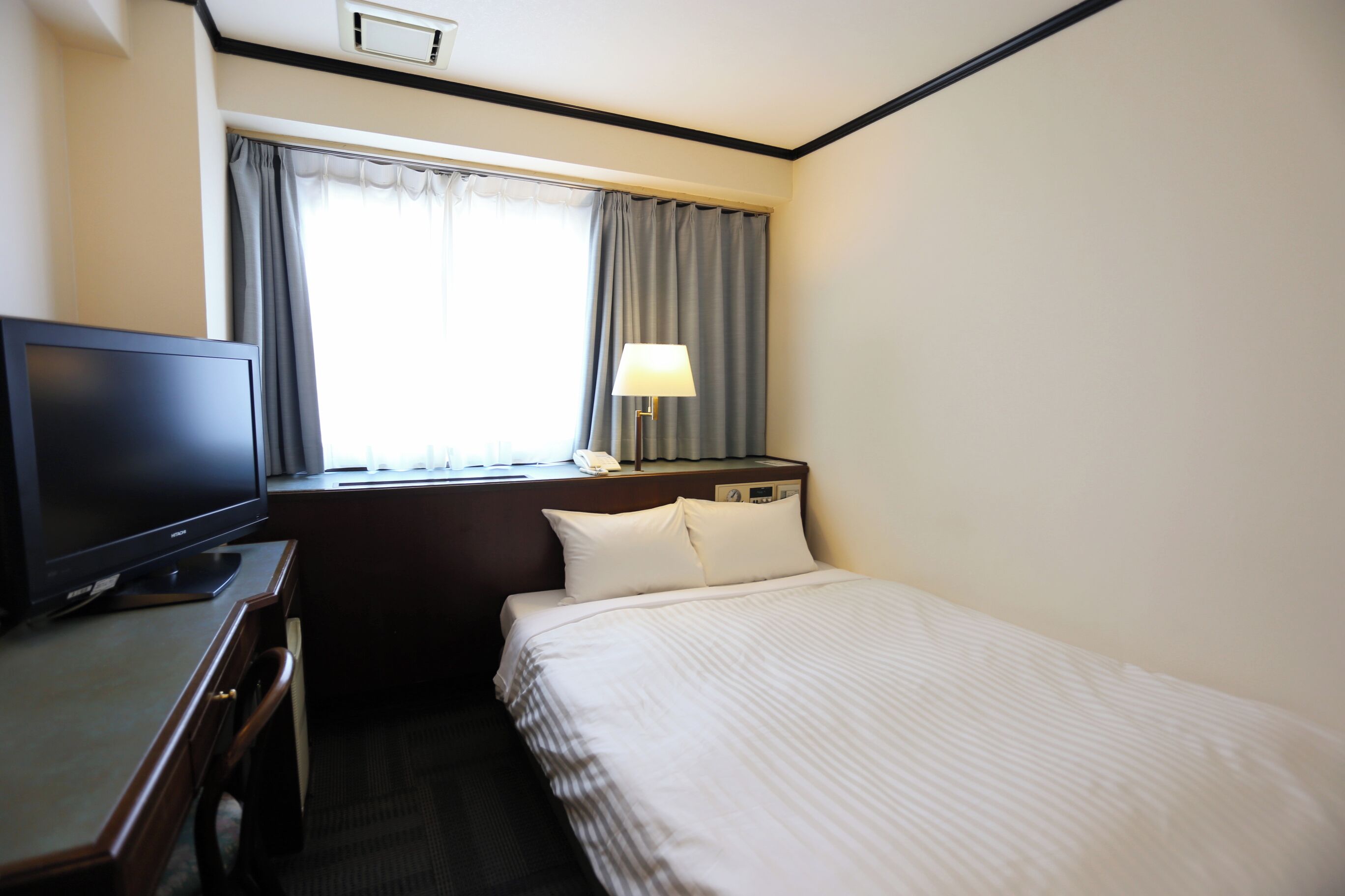 Habitación de hotel con cama, escritorio con televisor, lámpara y ventana con cortinas.