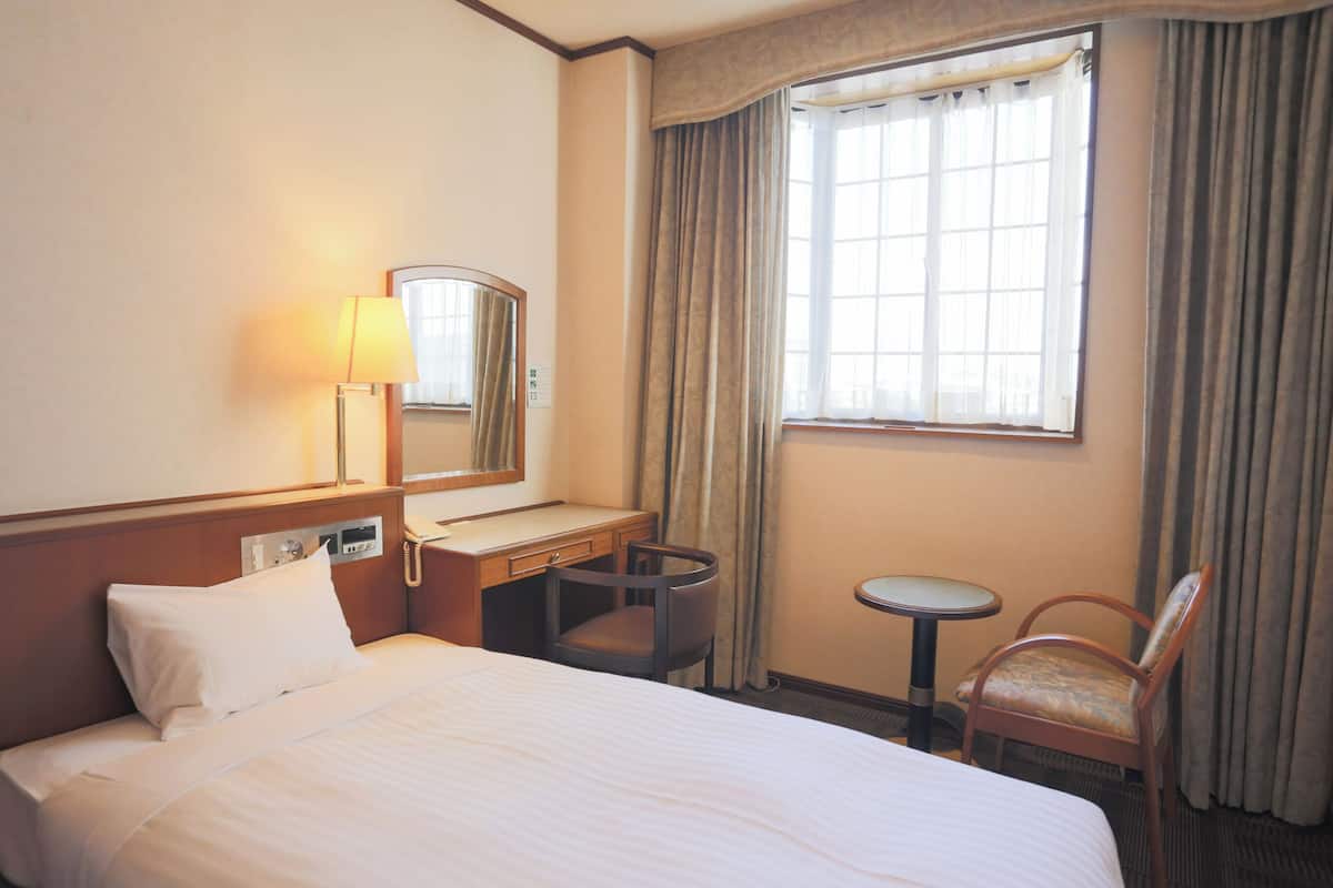 A hotel room with a bed, a desk, a chair, a small table, a lamp, a mirror, and a window with curtains.