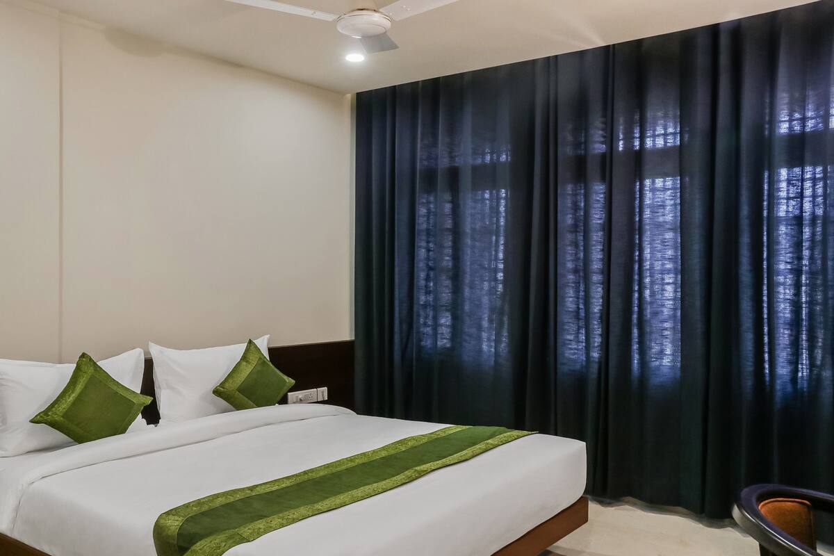 Una habitación de hotel con cama, ventilador de techo y ventana con cortinas oscuras.