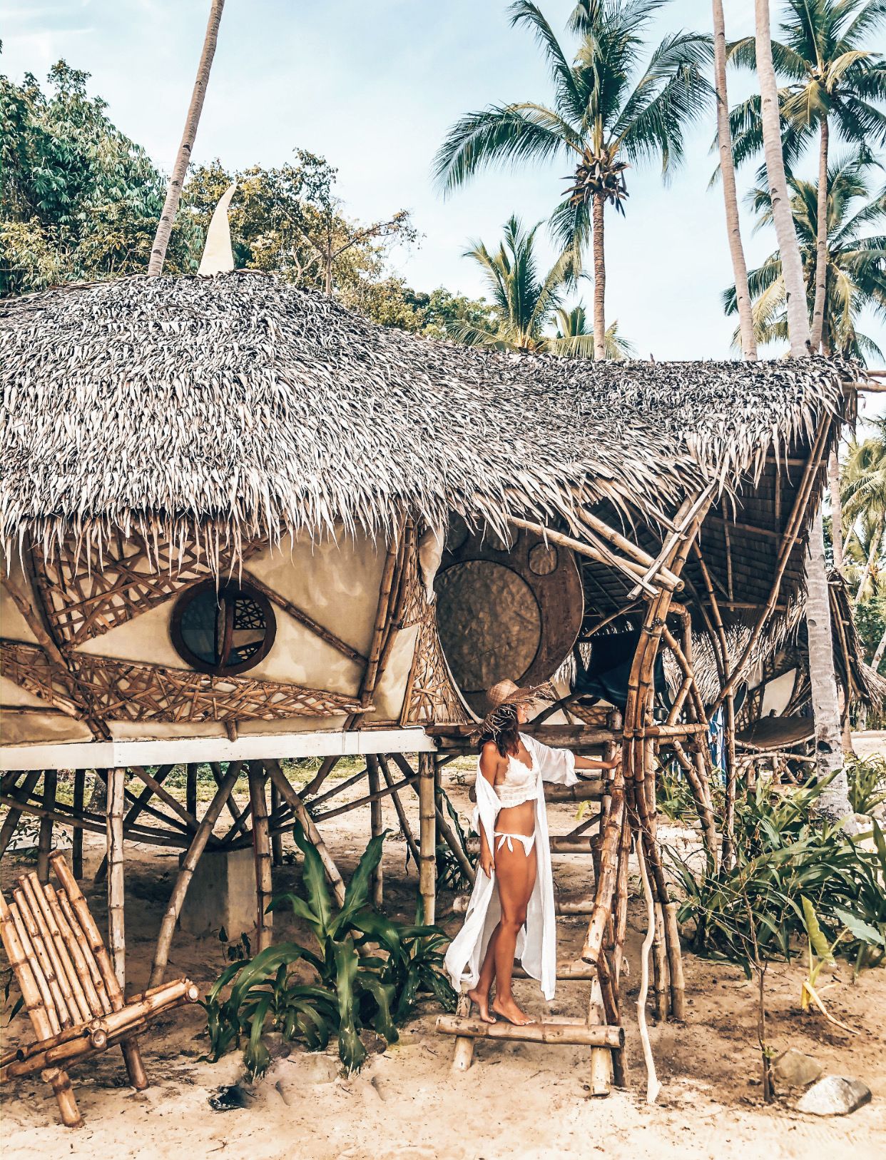 A thatched-roof hut with a circular window, surrounded by palm trees and a sandy ground.