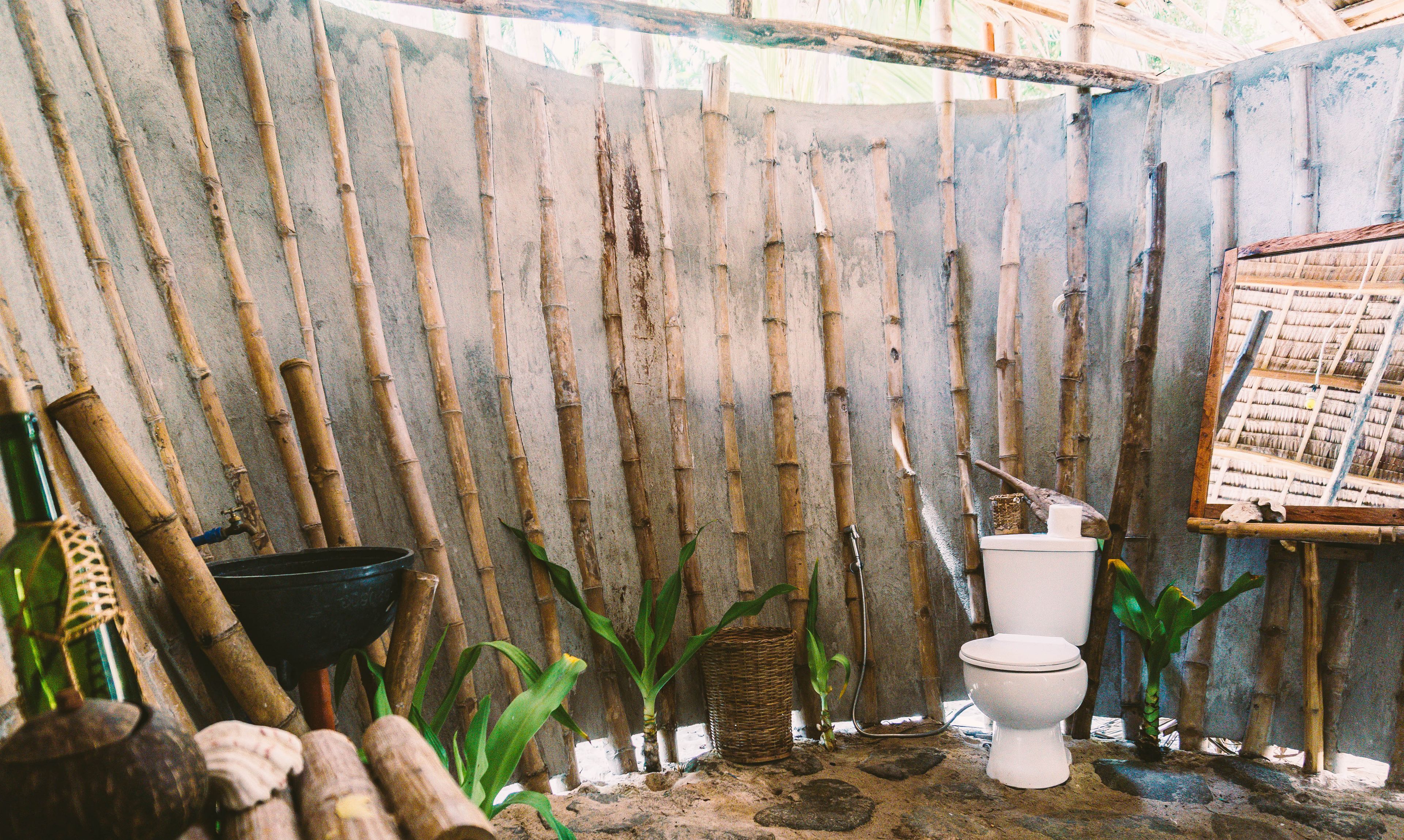 Un baño rústico con un muro de bambú, un inodoro blanco y un lavamanos negro pequeño.