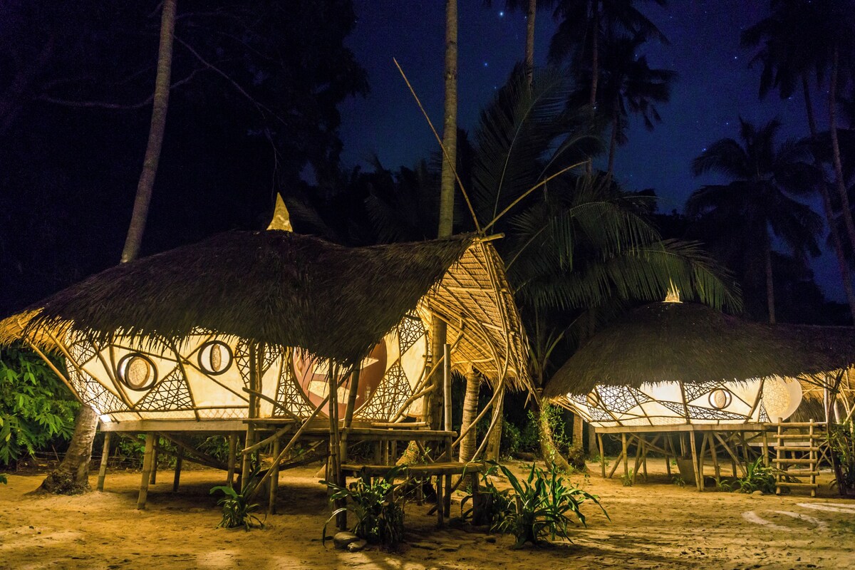 Cabanas com telhados de palha e janelas circulares, rodeadas por palmeiras, à noite.