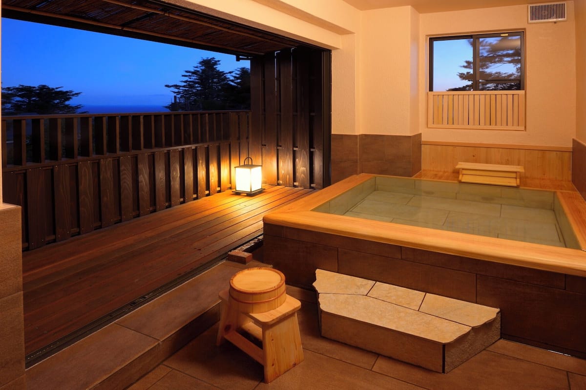 Un bain à remous avec une terrasse en bois, un petit tabouret et une fenêtre donnant sur une étendue d’eau.