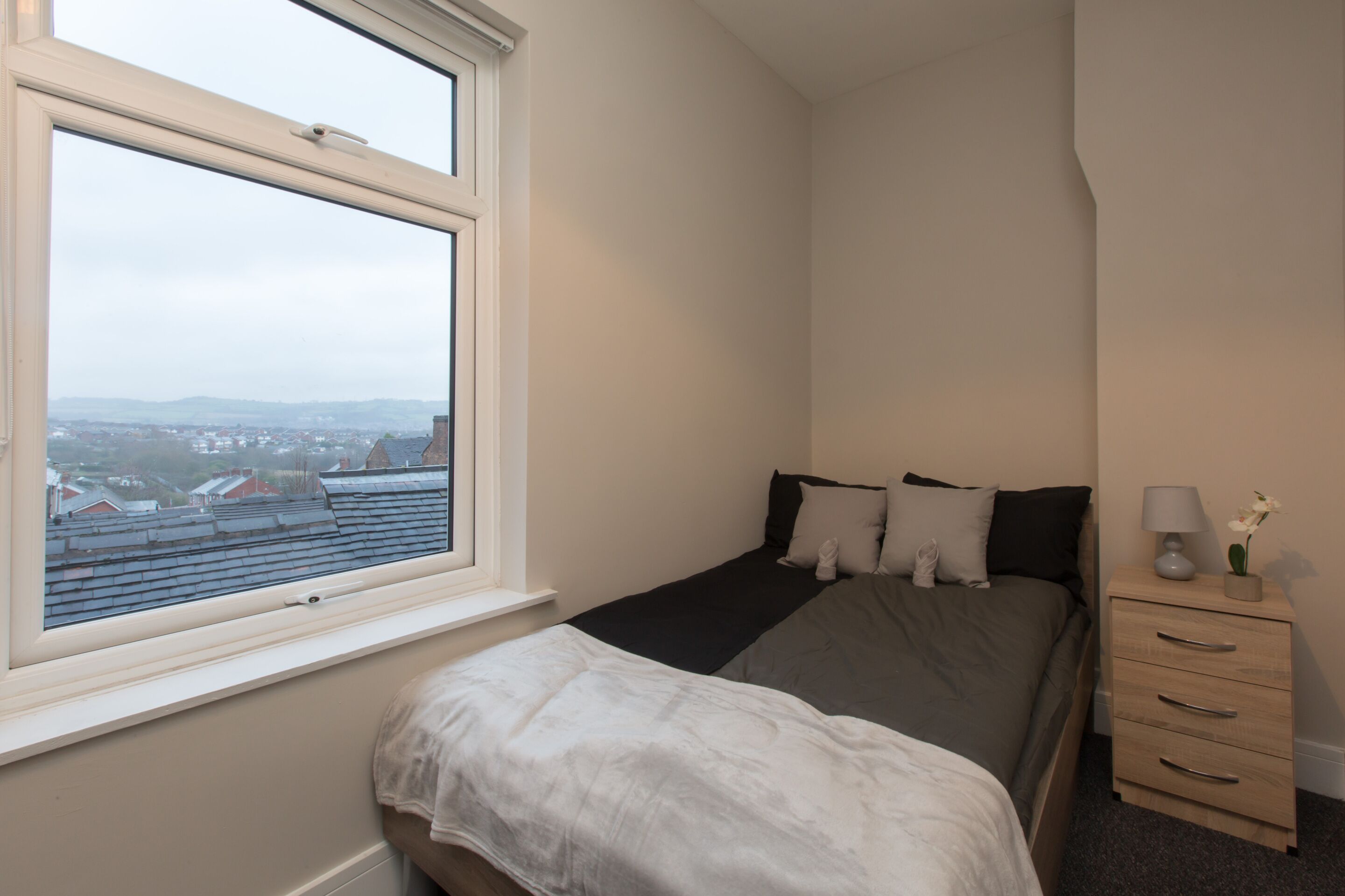 Un dormitorio con cama, mesita de noche, ventana y vistas a edificios.