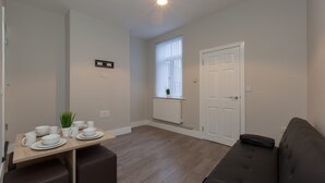 A modern living room with a sofa, a dining table set with white dishes, and a window with blinds. - Townhouse @ Rose Street Stoke (Stoke-on-Trent)