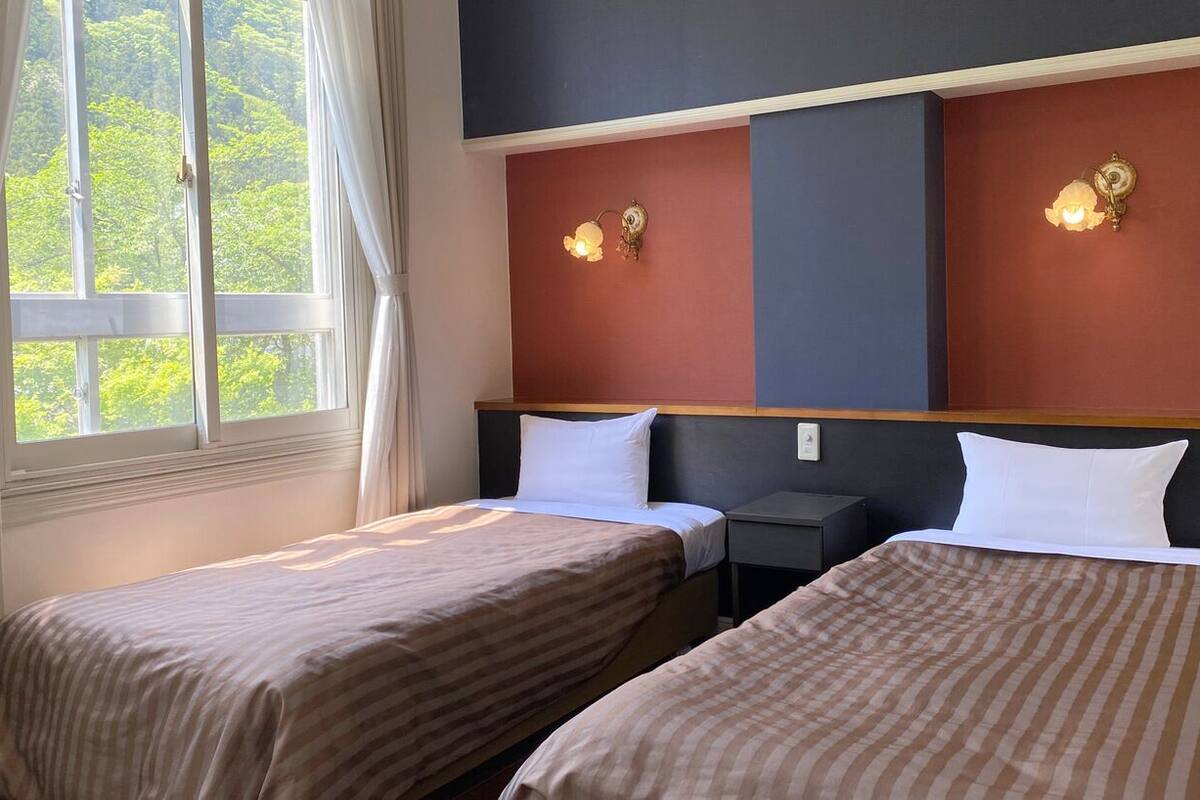 A hotel room with two beds, a window with curtains, and wall-mounted lamps.