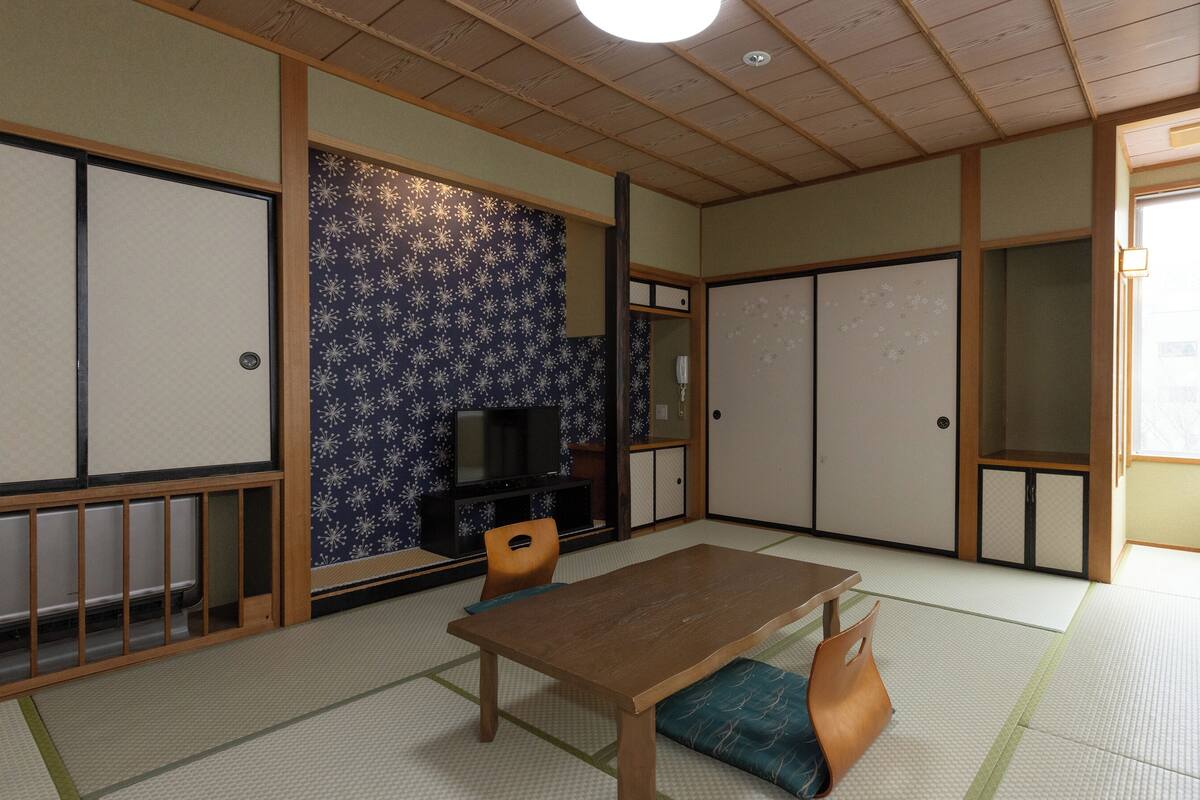 A traditional Japanese room with tatami flooring, a low table, a television, sliding doors, and a view through a window.