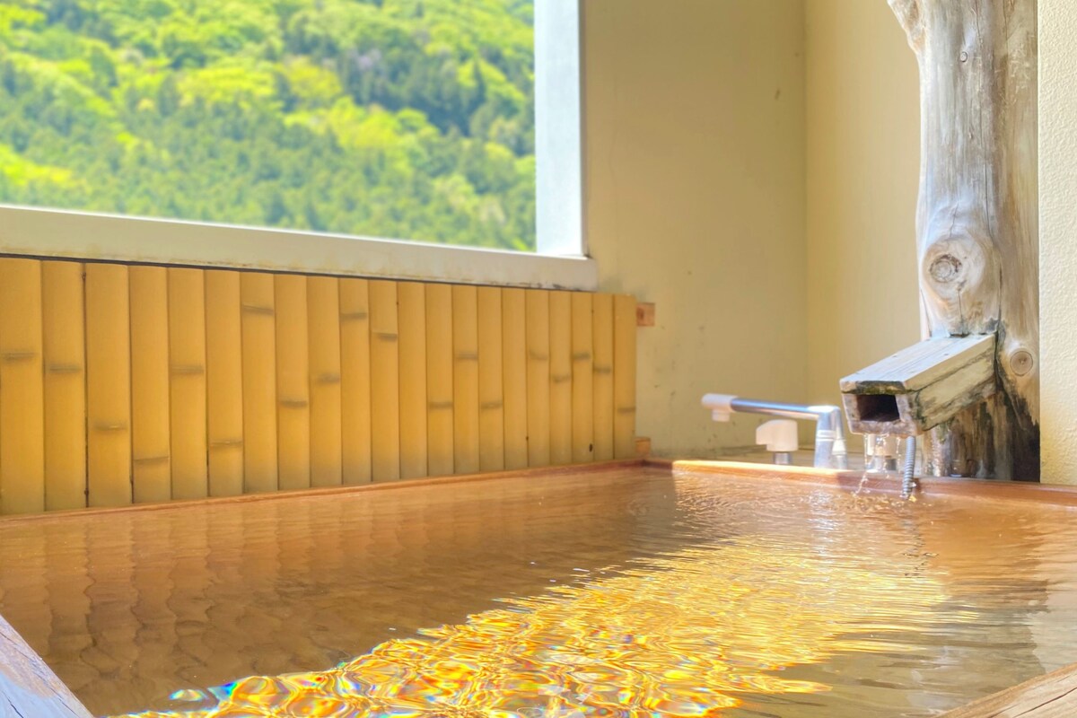 A wooden hot spring bath with a view of a forest through a window.