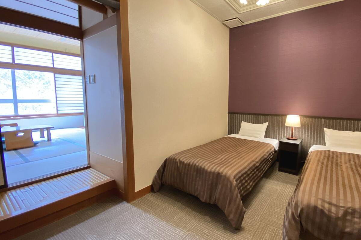 A hotel room with two beds, a nightstand with a lamp, a sliding door to a balcony, and a view of the outside.