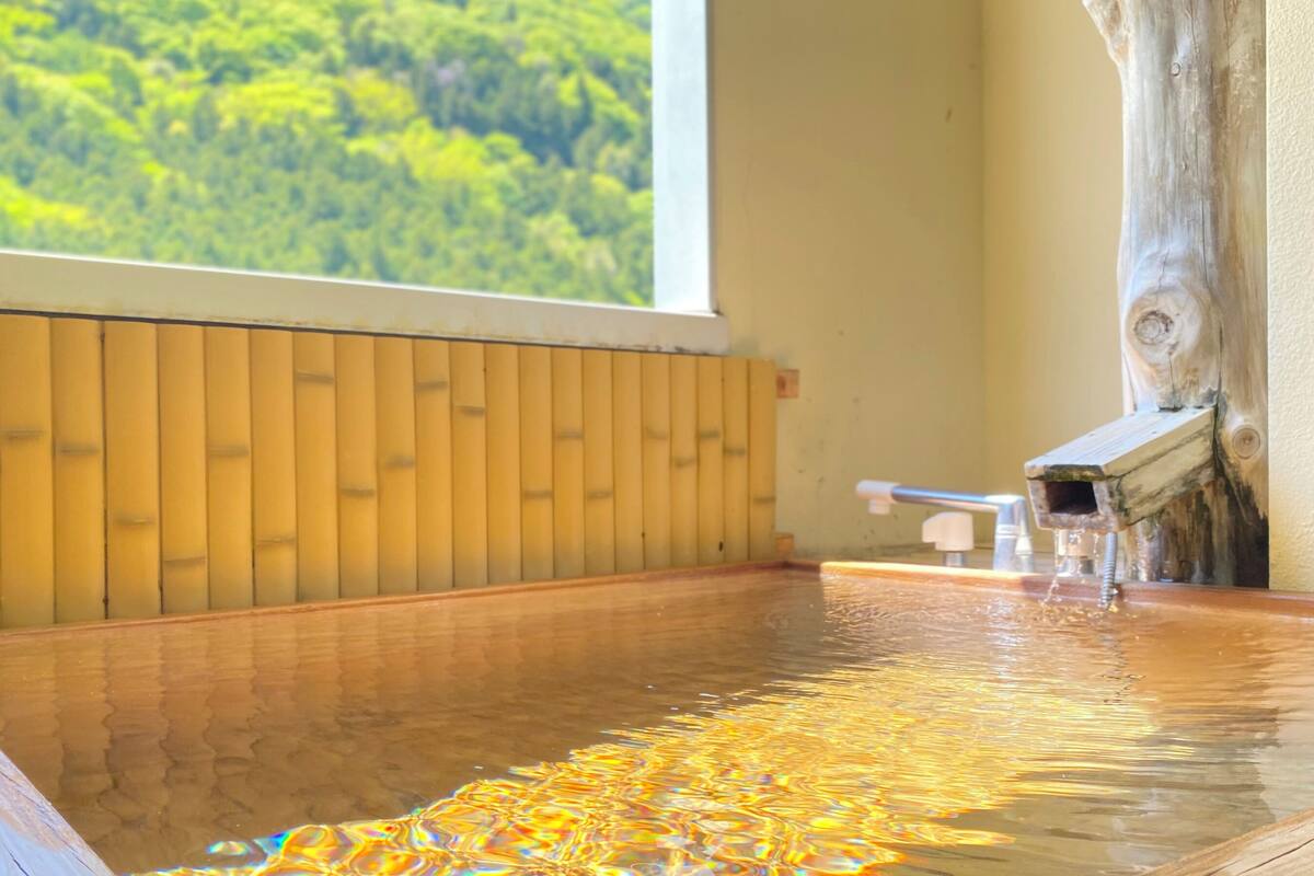 A wooden hot spring bath with a view of a forest through a window.