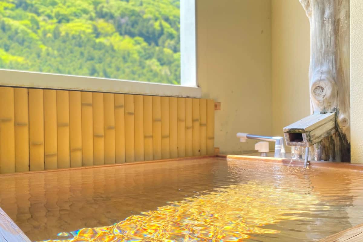 A wooden hot spring bath with a view of a forest through a window.
