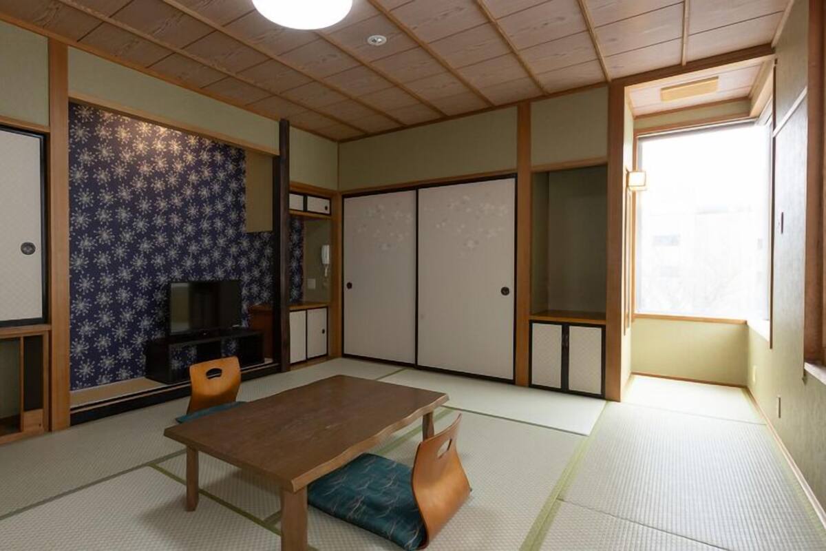 A traditional Japanese room with tatami flooring, a low wooden table, a television, sliding doors, and a view of the outside.