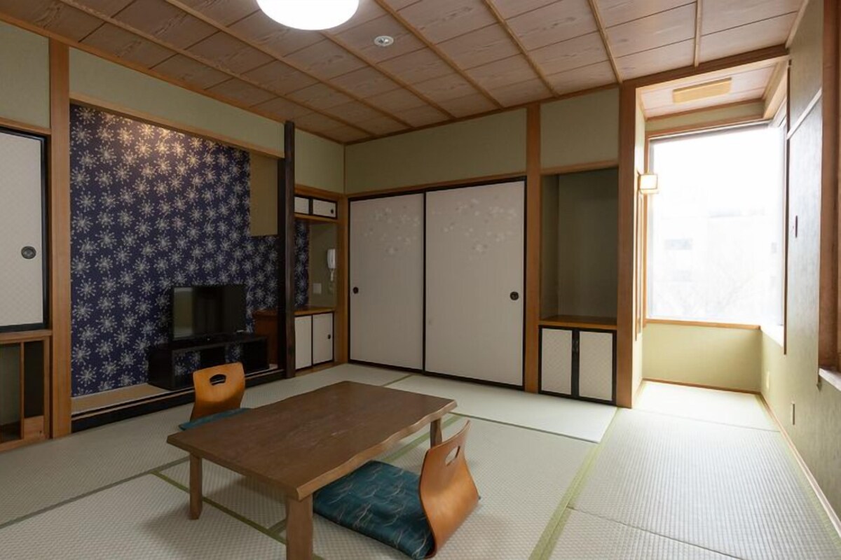 A traditional Japanese room with tatami flooring, a low wooden table, a television, sliding doors, and a view of the outside.