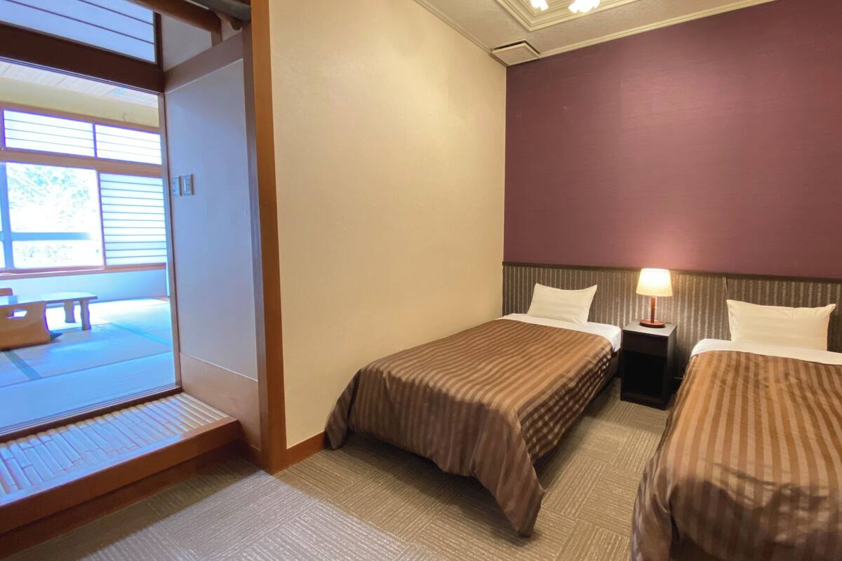 A hotel room with two beds, a small table, and a view of a deck area.