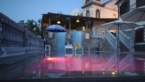Un bassin aux eaux calmes, bordé d’une balustrade en pierre, un parasol bleu et, au fond, un bâtiment doté d’un clocher.