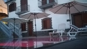 Un bâtiment à la façade blanche, doté de volets en bois et d’un balcon. On y trouve des chaises et des parasols à l’extérieur, ainsi qu’un escalier menant à l’entrée.