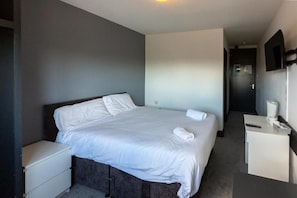 A hotel room with a single bed, a bedside table, a small desk with a chair, a mirror, and a door leading to another room.
