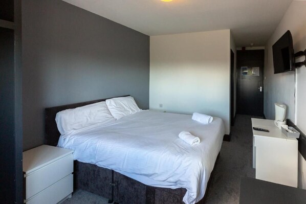 Habitación de hotel con una cama individual, una mesita de noche, un escritorio pequeño con una silla, un espejo y una puerta que da a otra habitación.