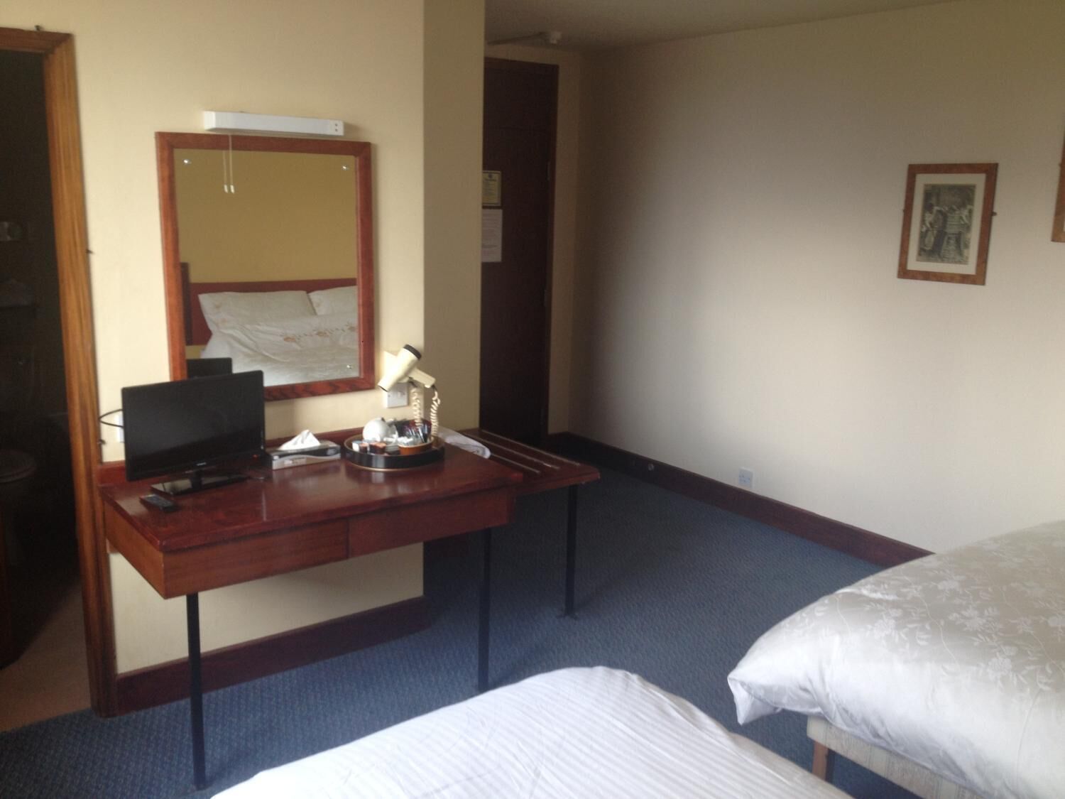 A hotel room with a wooden desk, a flat-screen TV, a lamp, a bed, and a large mirror.