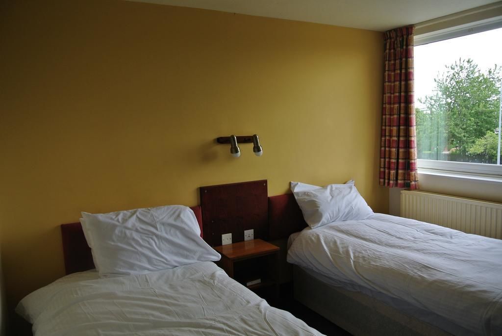A hotel room with two single beds, a wooden headboard, a nightstand, and a window with curtains.