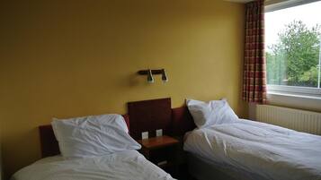 A hotel room with two single beds, a wooden headboard, a nightstand, and a window with curtains.