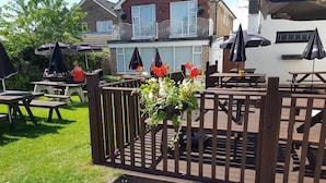 Een houten terras met picknicktafels en parasols, omringd door een hek en een huis op de achtergrond.