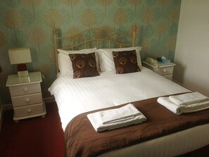 A neatly made bed with a brown bedspread and white pillows, a bedside table with a lamp, and a nightstand with a phone. - The Beverley Inn (Doncaster)