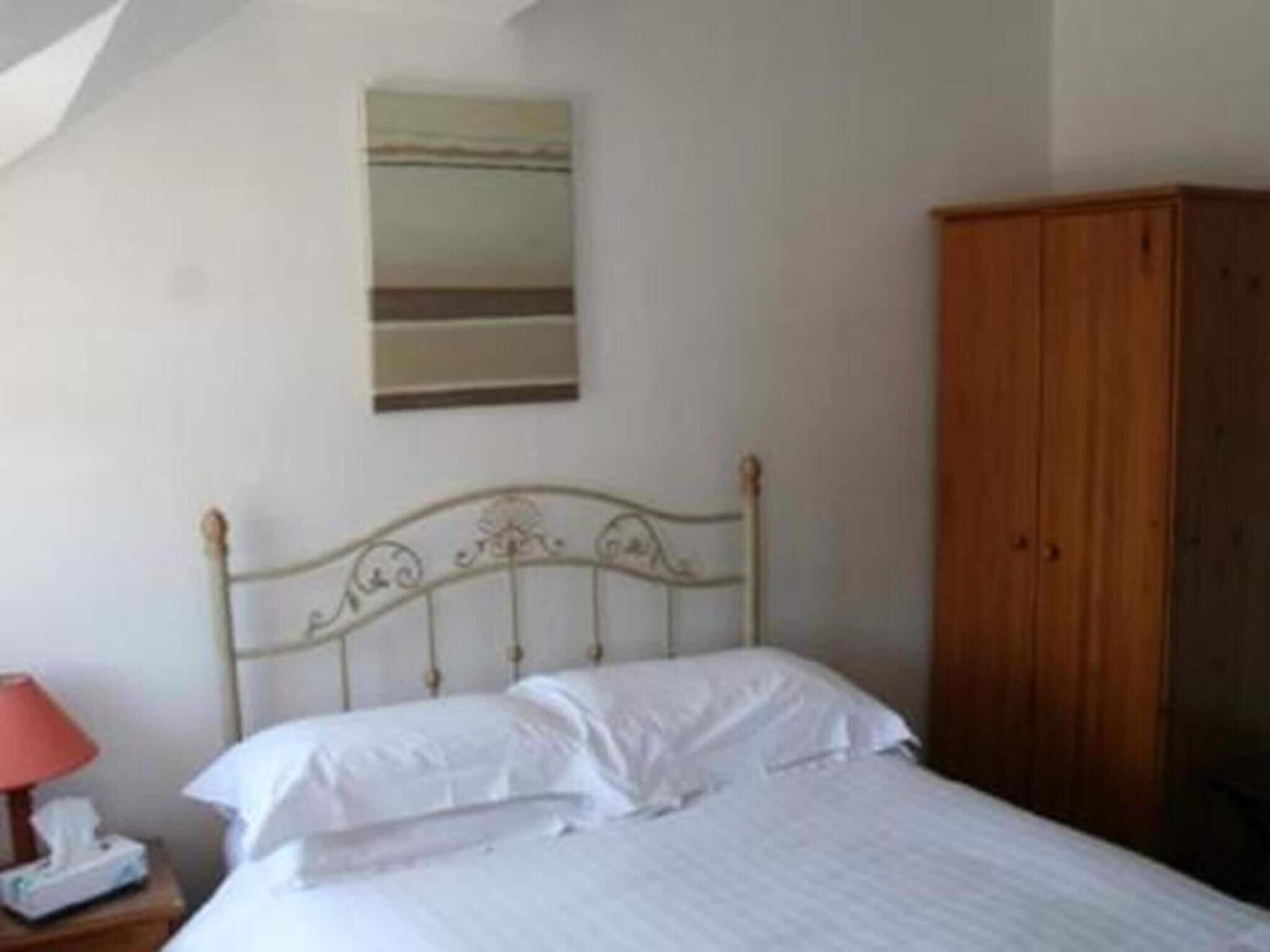 A bedroom with a metal bed frame, white bedding, a wooden wardrobe, and a bedside table with a lamp and a telephone.