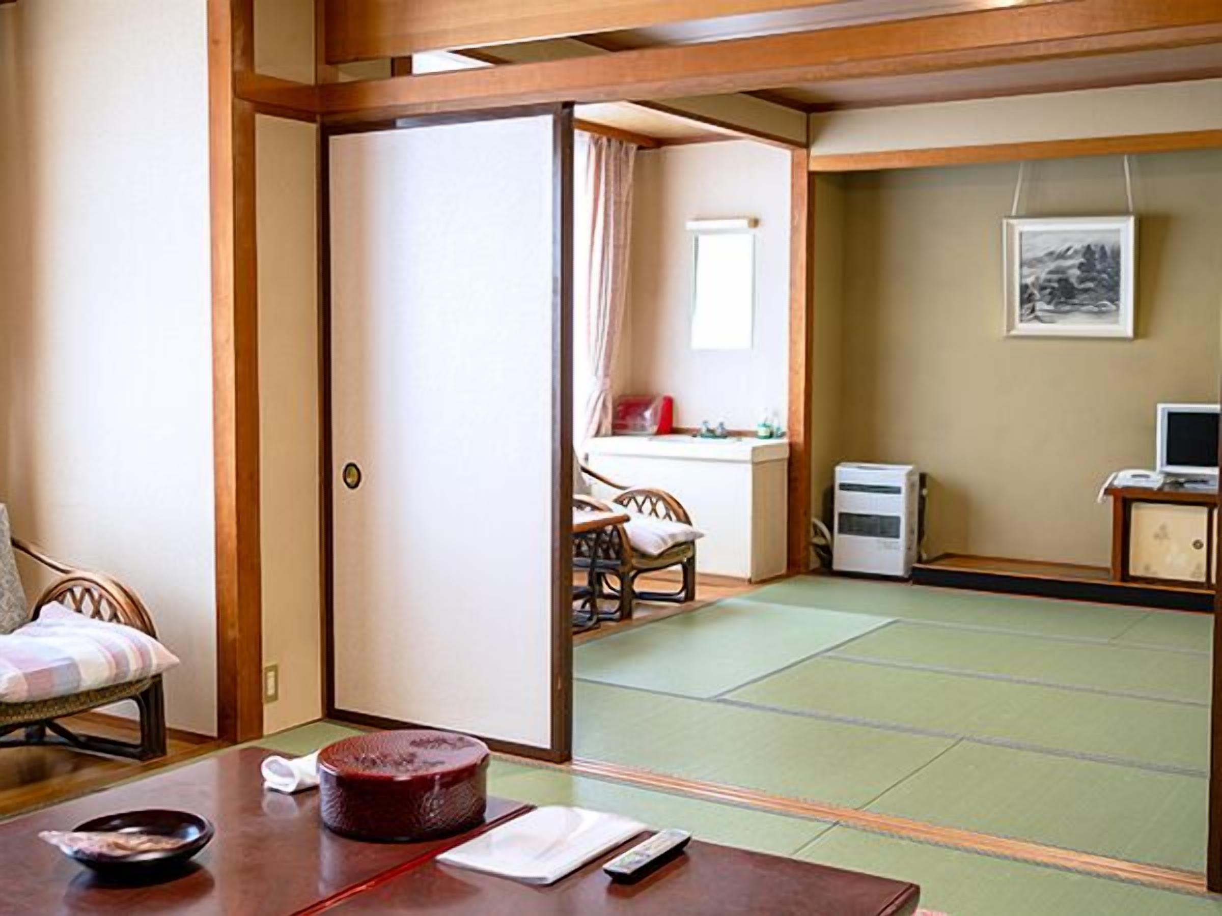A traditional Japanese room with tatami flooring, a tatami mat table, a low wooden desk, a chair, and a bed with a striped cushion.