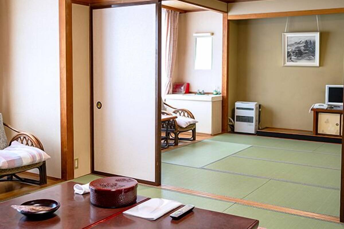 Ein traditioneller japanischer Raum mit Tatami-Böden, einem Tatami-Matten-Tisch, einem niedrigen Holzbüro, einem Stuhl und einem Bett mit gestreiftem Kissen.