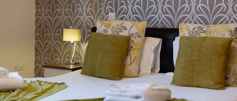 A hotel room with a neatly made bed, green and white floral pillows, a lamp on a bedside table, and a patterned wallpaper.