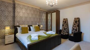 A neatly made bed with a green leaf decoration, a nightstand with a lamp, and two chairs with floral upholstery.