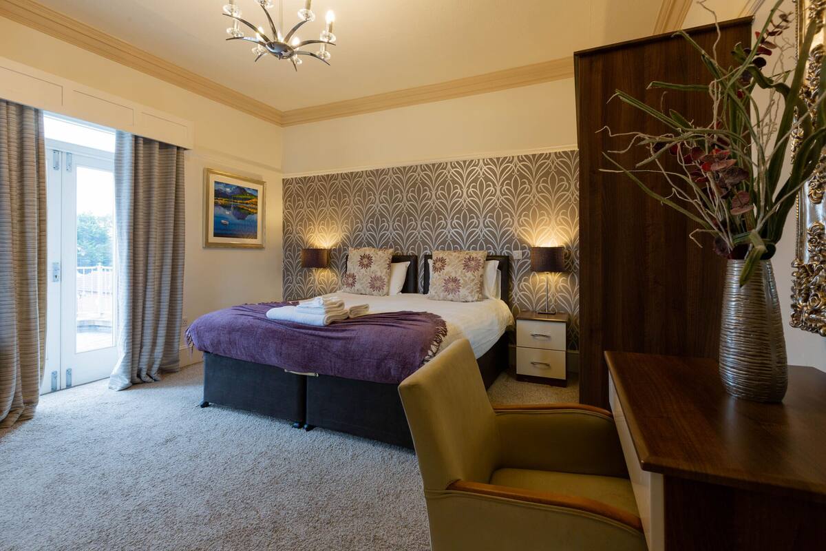 A hotel bedroom with a large bed, two bedside tables, a chair, a vase with flowers, and a painting on the wall.