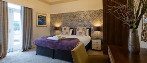 A hotel bedroom with a large bed, two bedside tables, a chair, a vase with flowers, and a painting on the wall.