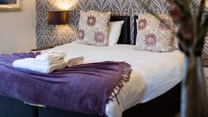 Ein ordentlich bezogenes Bett mit einer violetten Tagesdecke und gefalteten Handtüchern, einer Wandlampe und einem blumengemusterten Kopfteil.
