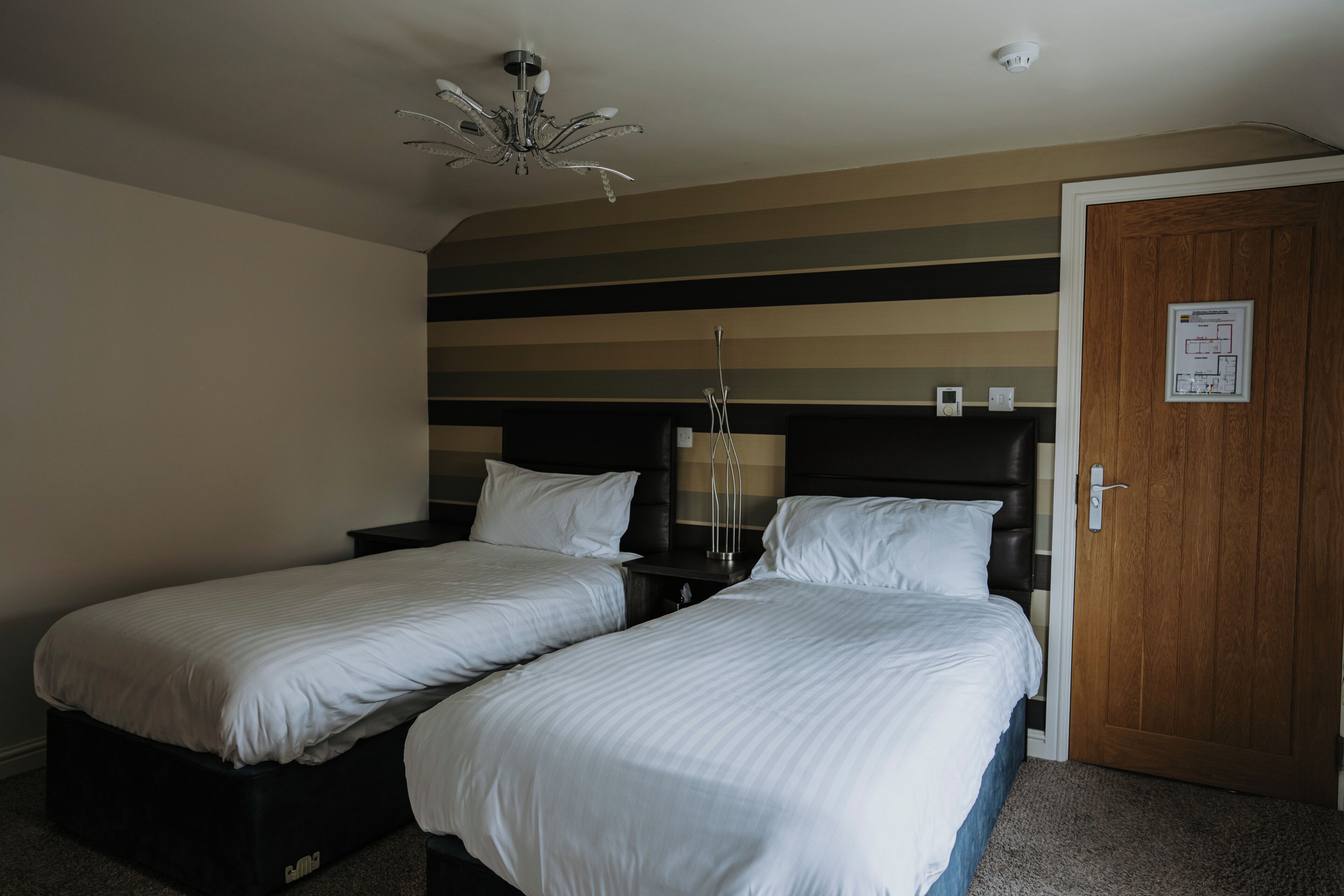 Habitación de hotel con dos camas, un candelabro, una puerta de madera e interruptor de luz en la pared.