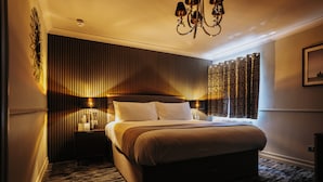 A hotel room with a large bed, a nightstand with a lamp, a wall-mounted light, and a window with patterned curtains. - The White Swan (Gainsborough)